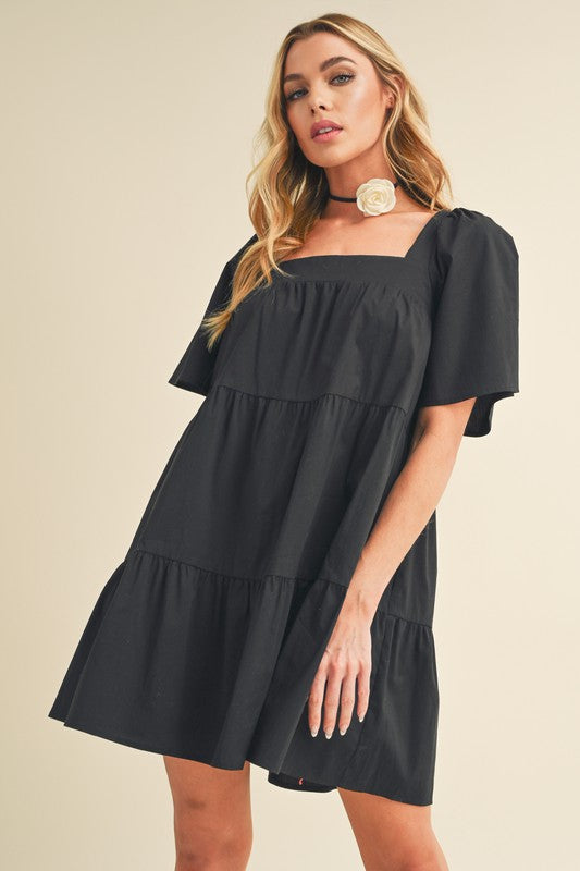 A woman wearing the Aemi + Co square neck half sleeve tiered dress by Jade Icarus, showcasing its stylish design and tiered fabric.