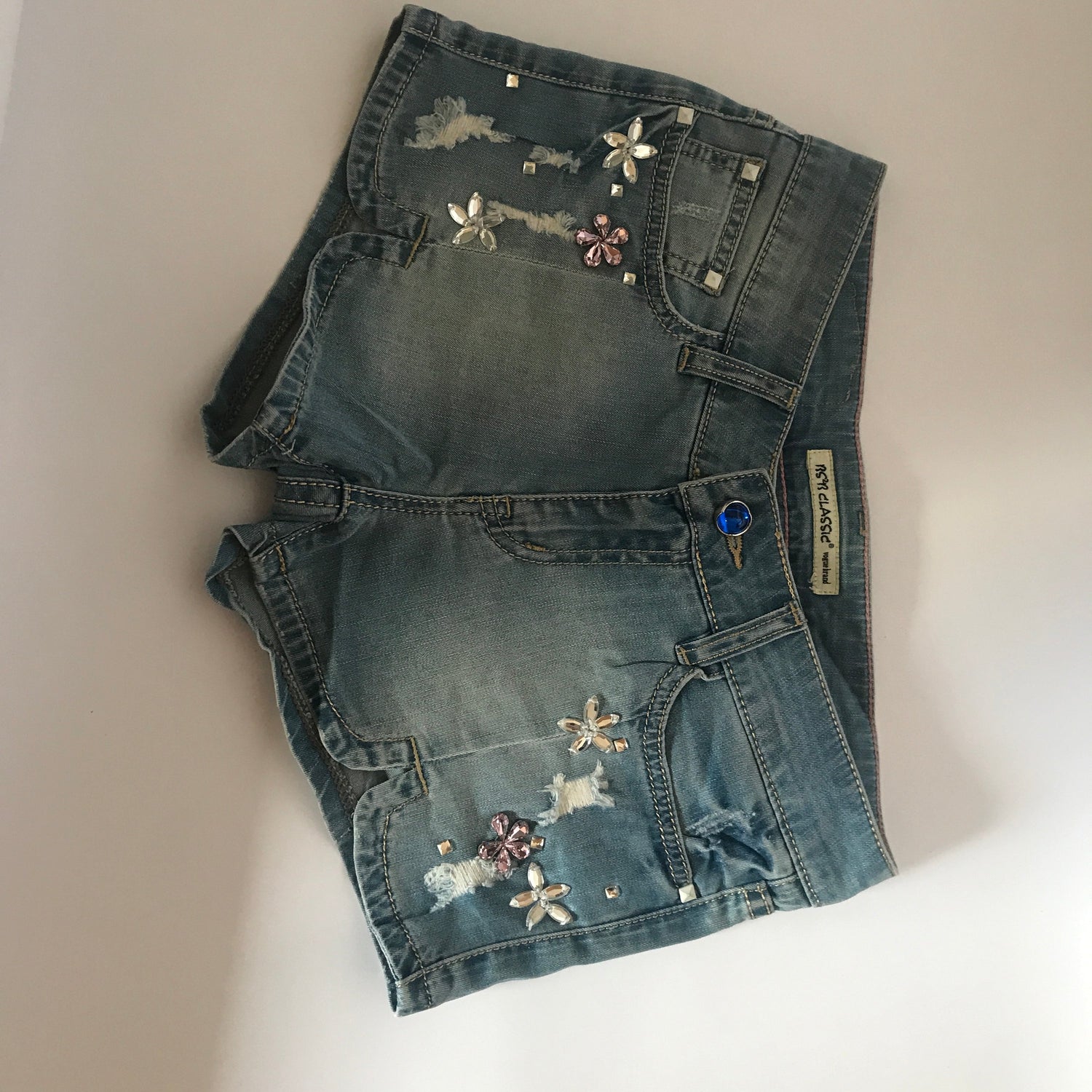 Blush Bella denim shorts decorated with flower-shaped rhinestones, showcasing stylish jewelry-inspired design.
