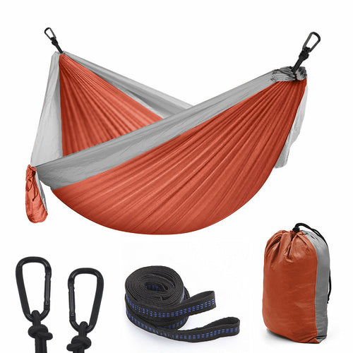 Maroon Asteria camping parachute hammock for outdoor survival, shown hanging in a garden with greenery and trees.