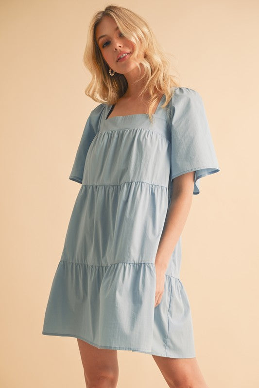 A white Aemi + Co square neck half sleeve tiered dress by Jade Icarus, displayed on a mannequin with a blurred background.